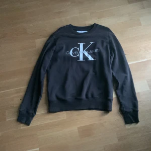 Svart sweatshirt från Calvin Klein Jeans - Svart sweatshirt från Calvin Klein Jeans i storlek XS. Tröjan har ett stort CK-tryck i vitt på bröstet och klassisk rund hals. Mjukt material och långa ärmar med ribbade muddar. Perfekt för en chill och stilren look.