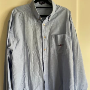 Gant skjorta ljusblå i bomull - Ljusblå skjorta från GANT med klassisk button-down krage och broderad logga på bröstfickan. Skjortan har långa ärmar, knappar framtill och är tillverkad i mjuk bomull. Perfekt för dig som gillar stilrena och tidlösa plagg. Kamerans röda sökare ger det lite bleka i mitten men skjortan är jämnt ljusblå.
