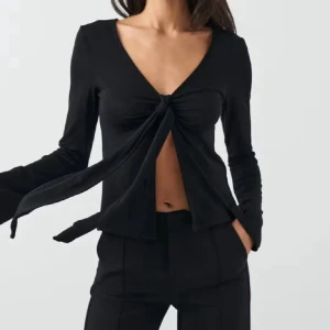 Svart långärmad knytblus - Svart långärmad topp med djup v-ringning och knyt framtill som ger en öppen detalj över magen. Toppens material är mjukt och stretchigt, vilket gör den bekväm och smickrande. Perfekt att styla med både jeans och kjol för en trendig look.