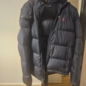 Mörkblå dunjacka Polo Ralph Lauren - Snygg mörkblå dunjacka från Polo Ralph Lauren med röd logga på bröstet. Jackan har quiltad design, tvåvägsdragkedja och flera fickor med dragkedja. Perfekt för kalla dagar och riktigt skön att bära.