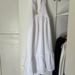 Vit somrig klänning med volang - Luftig vit klänning med smock upptill och volang nertill. Klänningen är ärmlös med öppen rygg och knyt i nacken. Perfekt för varma dagar med sin flowiga passform och tunna material. 💞💞