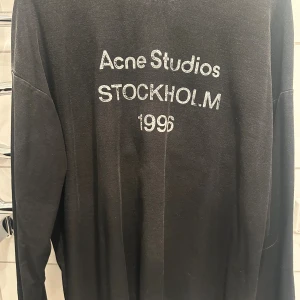 Svart långärmad tröja Acne Studios - Svart långärmad tröja från Acne Studios med tryck i vitt på bröstet: 'Acne Studios STOCKHOLM 1996'. Enkel och stilren design med rund hals. Perfekt för dig som gillar minimalistiskt mode och vill ha något från ett ikoniskt svenskt märke.