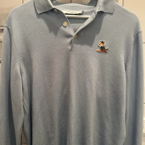 Ljusblå pikétröja Maison Kitsuné - Ljusblå långärmad pikétröja från Maison Kitsuné med klassisk krage, knäppning med tre knappar och broderad rävpatch på bröstet. Tröjan är i mjuk bomull och har en clean, stilren look som passar perfekt till jeans eller chinos.