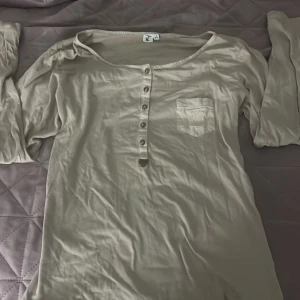 Beige långärmad topp från H&M - Säljer en beige långärmad topp från H&M med rund halsringning, knappslå framtill och liten bröstficka med broderad detalj. Tunt och mjukt material som känns skönt mot huden. Perfekt för lager-på-lager och enkel att styla.