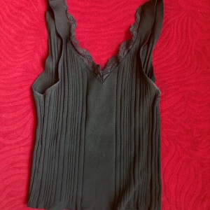 Svart ribbad linnetopp med spets - Svart ribbad linnetopp med breda axelband och v-ringning dekorerad med spets. Toppen är ärmlös och har en figurnära passform. Perfekt att styla med jeans eller kjol för en snygg look.