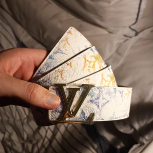 Vit Louis Vuitton bälte med färgstänk - Säljer ett snyggt vitt Louis Vuitton-bälte med färgstänk i blått, gult och orange. Bältet har en ikonisk LV-spänne i guldmetall och detaljerad struktur i skinn. Perfekt accessoar för att lyfta din outfit med en exklusiv touch.
