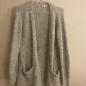 Ljusgrå stickad kofta med fickor - Mysig ljusgrå stickad kofta med långa ärmar och två stora fickor framtill. Koftan har en öppen design utan knappar eller dragkedja och är perfekt att slänga på sig när det är kyligt. Enkel och stilren look som passar till allt.