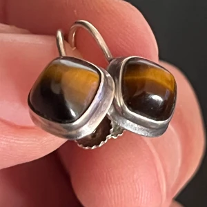 Örhängen i silver med tigeröga - Coola örhängen i äkta silver med fyrkantiga cabochoner av tigeröga. Stenarna har en snygg mix av bruna och gyllene toner som skiftar i ljuset. Skruvstängning för extra säkerhet och retrovibe. Perfekt accessoar för dig som gillar unika detaljer.