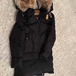 Svart Parajumpers pufferjacka XS - Säljer en svart pufferjacka från Parajumpers i storlek XS. Jackan har en mysig fuskpäls på huvan, patch på ärmen och coola detaljer som dragkedja och knappar. Perfekt för kalla dagar med sin långa modell och vadderade känsla.