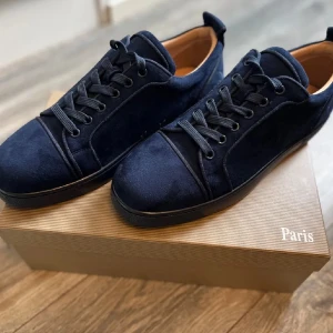 Mörkblå sneakers Christian Louboutin - Snygga mörkblå sneakers från Christian Louboutin i mocka med klassisk röd sula. Skorna har rund tå, svarta snören och diskret broderad logga på hälen. Insidan är ljusbeige och sulan är platt. Perfekta för dig som vill ha en lyxig och stilren look.