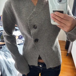 Grå kofta från Zara med asymmetrisk knäppning - Mysig grå kofta från Zara med unik asymmetrisk knäppning och två stora knappar framtill. Koftan har lång ärm och är tillverkad i ett mjukt, stickat material som känns riktigt skönt mot huden. Lite nopprig 
