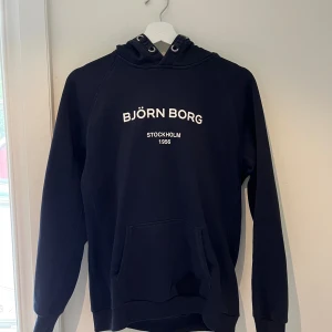 Blå hoodie från Björn Borg - Blå hoodie från Björn Borg med vit text på bröstet. Klassisk känguruficka framtill, huva med snörning och mjukt material. Perfekt för chill dagar eller när du vill ha en enkel och snygg look.