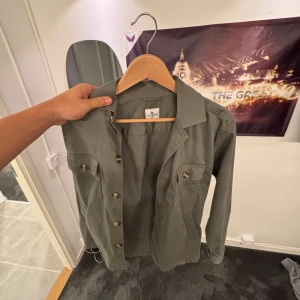 Grön overshirt från John Henric - Stilren grön overshirt från John Henric med klassisk krage, fyra fickor framtill och snygga knappar. Jackan är tillverkad i bomull och har en rak passform med långa ärmar. Perfekt att slänga över en t-shirt för en clean look. Använd fåtal gånger. Jag är 180 och passar perfekt 