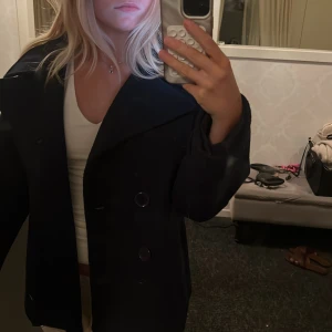 Mörkblå skepparkavaj från Design by Si XS/S - Stilren mörkblå skepparkavaj från Design by Si i storlek XS/S. Jackan har bred krage, dubbelknäppning med stora knappar och en rak, boxig passform. Perfekt för dig som gillar klassiska vibbar med en modern twist.