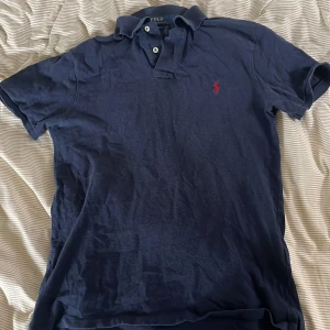 Mörkblå Polo Ralph Lauren pike - Snygg mörkblå t-shirt från Polo Ralph Lauren i slim fit med klassisk krage och två knappar. Ikoniska röda logon broderad på bröstet. Tillverkad i mjuk bomull som är skön mot huden. Perfekt för dig som gillar stilrena och tidlösa plagg.