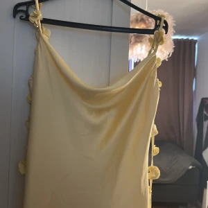 Ljusgul långklänning med blommor - Säljer en ljusgul långklänning med smala axelband och dekorativa blommor längs banden och sidan. Klänningen har en mjuk, draperad urringning och är tillverkad i ett glansigt, slätt material som ger en lyxig känsla. Perfekt för dig som vill sticka ut med en feminin och elegant look.Den är helt ny har aldrig använt den och prislappen är fortfarande på
