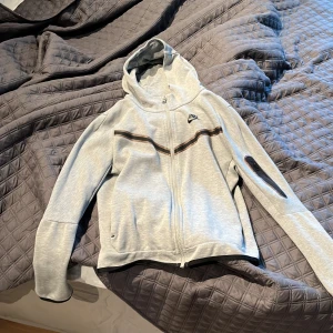Grå hoodie från Nike med dragkedja - Snygg ljusgrå hoodie från Nike med hel dragkedja och huva. Tröjan har svarta och bruna ränder över bröstet och en liten ficka med dragkedja på ena ärmen. Perfekt för chill eller träning, klassisk Nike-logga på bröstet.