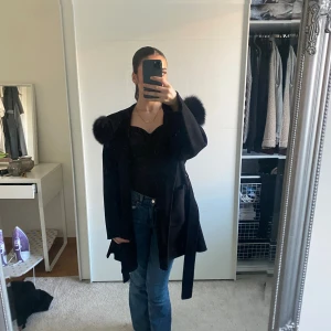 Svart kappa med äkta päls - Säljer en svart kappa med äkta päls på huvan. Jackan har ett bälte i midjan och två stora fickor framtill. Modellen är loose och har långa ärmar, perfekt för dig som gillar statement-plagg med extra attityd. Pasar till både höst,vår och vinter. Den har använts 2 gånger! Den är i storlek S men passar folk i Xs oxå.