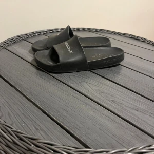 Svarta sandaler från Calvin Klein - Svarta sandaler från Calvin Klein med enkel och stilren design. Tillverkade i syntetmaterial med bred rem över foten och logga på ovansidan. Perfekta för sommaren eller att slänga på sig till stranden. Platt sula och öppen tå för en avslappnad vibe. Den var några söndriga som bra syns när man sträcker på tofflan annars syns den inte jätte tydligt och är jätte bra