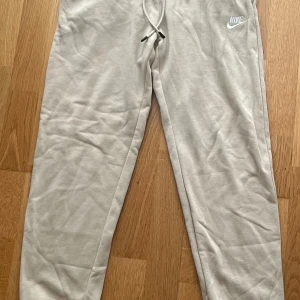 Beige mjukisbyxor från Nike - Säljer ett par beige mjukisbyxor från Nike med dragsko i midjan och klassisk logga på ena benet. Byxorna har en avslappnad passform och är tillverkade i mjukt bomullsmaterial, perfekta för chill dagar eller när du vill vara bekväm.