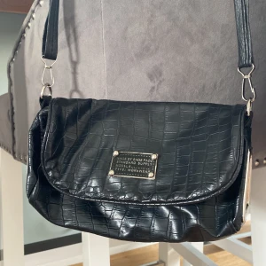 Svart axelväska med croco-mönster - Snygg svart axelväska i croco-mönstrat skinnimitation med silverfärgade metalldetaljer och justerbar axelrem. Väskan har lock med magnetknäppning och ett mönstrat innerfoder med dragkedjefack. Perfekt för dig som vill ha en stilren och trendig väska.