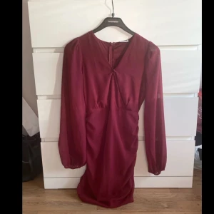 Vinröd långärmad klänning med v-ringning - Snygg vinröd klänning med långa ärmar och v-ringning framtill. Klänningen har en lätt rynkad detalj vid bysten och är tillverkad i ett mjukt, flowy material som ger en elegant känsla. Perfekt för dig som vill sticka ut med en djup färg och stilren design. Nederdelen av v-ringningen är sydd då det var en för djup ringning. 