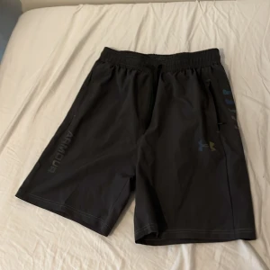 Svarta träningsshorts från Under Armour - Säljer ett par svarta träningsshorts från Under Armour i loose fit. Shortsen har elastisk midja med snörning, reflekterande logga och text på sidan. Materialet är lätt polyester som andas, perfekt för gym eller sport. Snyggt och stilrent med coola detaljer.