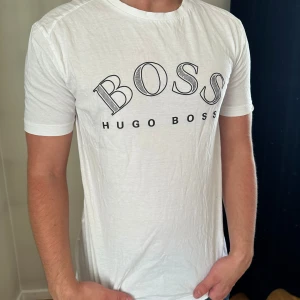 Vit Hugo Boss t-shirt med tryck - Vit t-shirt från Hugo Boss med stort BOSS-tryck i svart på bröstet. Klassisk rund hals och korta ärmar. Materialet är mjuk bomull som känns skönt mot huden. Passformen är normal och t-shirten är enkel att matcha med jeans eller shorts.
