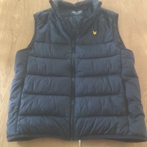 Svart dunväst från Lyle & Scott - Svart dunväst från Lyle & Scott med gul broderad logga på bröstet. Västen har quiltad design, hög krage och dragkedja framtill. Perfekt för lager-på-lager och har en clean, sportig look som funkar till många stilar.