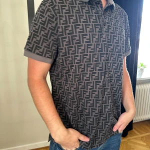 Grå Fendi t-shirt med FF-mönster - Säljer en grå t-shirt från Fendi med det ikoniska FF-mönstret över hela plagget. T-shirten har korta ärmar, krage och knappar vid halsen. Materialet är mjukt och känns som bomull, perfekt för dig som gillar designer och vill sticka ut.