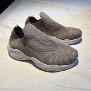 Grå slip-on sneakers från WUUD - Gråa slip-on sneakers från WUUD med vit, chunky sula. Ovandelen är i stickad syntet med meshstruktur som ger en sportig vibe. Skorna har rund tå och elastisk öppning för enkel påtagning. Perfekta för dig som gillar minimalistisk och clean stil.