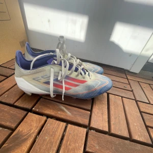 Adidas blå och röda fotbollsskor - Säljer ett par Adidas fotbollsskor med snygg design i blått, vitt och rött. Skorna har snörning, platt sula med dobbar och är tillverkade i syntetmaterial. Perfekta för dig som vill sticka ut på planen med stilrena och sportiga skor.