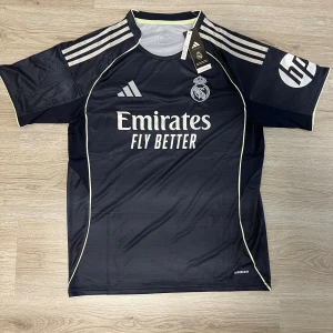 Real Madrid Adidas borta fotbollströja svart L - Snygg Real Madrid fotbollströja från Adidas i svart med tunna horisontella ränder. Tröjan har korta ärmar, vita detaljer och klubbmärke samt sponsortryck på bröstet. Tillverkad i lätt och ventilerande polyester med klassiska Adidas-ränder på axlarna.