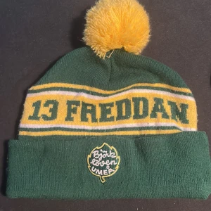 Björklöven mössa med tofs och Freddan-tryck - Grön och gul stickad mössa med stor gul tofs på toppen. Mössan har texten '13 Freddan' i grönt och gult samt ett broderat Björklöven Umeå-märke framtill. Perfekt för dig som vill visa ditt stöd för laget och hålla dig varm på matchen.