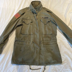 Olivgrön parkajacka från Ralph Lauren - Snygg olivgrön parkajacka från Ralph Lauren med patchad amerikansk flagga på axeln och broderad logga på bröstet. Jackan har flera stora fickor, hög krage, snörning i midjan och axelklaffar för en cool militärinspirerad look.