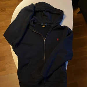 Marinblå hoodie från Polo Ralph Lauren - Säljer en marinblå hoodie från Polo Ralph Lauren med dragkedja och huva. Klassisk röd logga broderad på bröstet, känguruficka framtill och långa ärmar. Perfekt för dig som gillar stilren och bekväm streetwear.