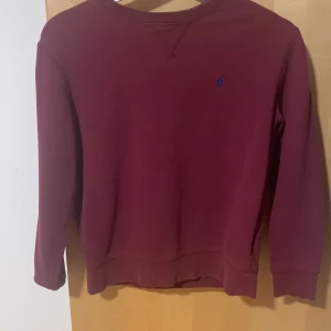 Vinröd tröja från Polo Ralph Lauren - Vinröd långärmad tröja från Polo Ralph Lauren med klassisk rund halsringning och blå broderad logga på bröstet. Tröjan är i mjuk bomull och har en enkel, stilren design som passar perfekt till jeans eller chinos.