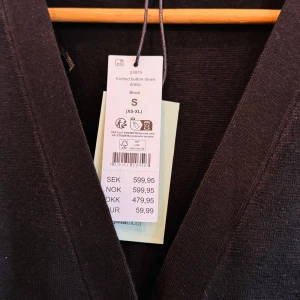 Svart stickad långklänning S - Svart stickad långklänning med v-ringning och knappar hela vägen framtill. Klänningen har lång ärm och är gjord i ett mjukt stickat material som sitter snyggt på kroppen. Perfekt för dig som gillar enkel och stilren design.