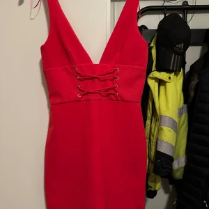 Röd kortklänning med snörning strl: Xs - Snygg röd kortklänning med djup v-ringning och cool snörningsdetalj framtill i midjan. Klänningen är ärmlös och har en figurnära passform som framhäver siluetten. Perfekt för dig som vill sticka ut med en enkel men ändå iögonfallande look.