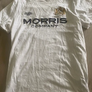 Vit t-shirt från Morris Company - Vit t-shirt från Morris Company med stor logga och broderat emblem på bröstet. Klassisk rund hals och korta ärmar. Skön och stilren modell i bomull, perfekt till jeans eller shorts. Snygg detalj med upphöjd text och guldbroderi.
