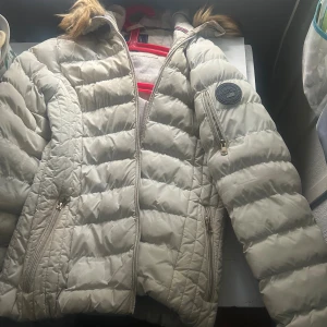 Beige pufferjacka Vinson Polo Club XS - Snygg beige pufferjacka från Vinson Polo Club med huva och fuskpälsdetalj. Jackan har quiltad design, dragkedjor på fickor och ärmar samt en patch på ena ärmen. Perfekt för kyliga dagar och riktigt skön att bära. Inga defekter. Pris kan diskuteras 