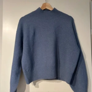 Blå stickad tröja med hög krage - Mysig blå stickad tröja med hög ribbad krage och breda muddar. Tröjan har en loose passform och långa ärmar, perfekt för kyliga dagar. Enkel och stilren design som är lätt att matcha med olika outfits.
