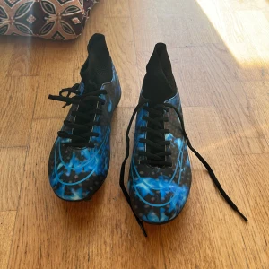 Blå-svarta fotbollsskor nike - Säljer ett par riktigt snygga fotbollsskor i storlek 36 med svart bas och blått mönster som ser ut som eldflammor. Skorna har snörning och en smal, sportig form med rund tå. Perfekta för dig som vill sticka ut på planen!