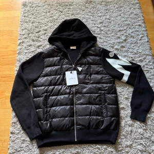 Moncler cardigan - Storlek L/Xl. Svart hoodie från Moncler med quiltad, glansig front i syntet och stickade ärmar. Vit zigzag-detalj på ena ärmen och Moncler-logga på axeln. Huva med snörning och hel dragkedja framtill. Perfekt för kyliga dagar.