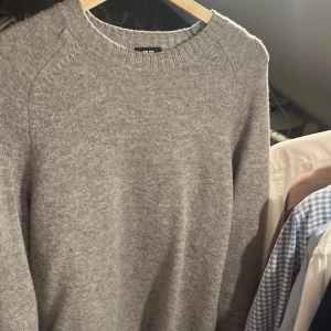 Grå stickad tröja med rund hals - Mysig grå stickad tröja från Uniqlo med rund halsringning och långa ärmar. Perfekt för lager-på-lager och enkel att matcha med jeans eller kjol. Tröjan har en klassisk passform och ribbade muddar vid hals, ärmslut och nederkant.