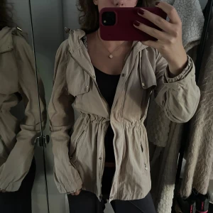 Beige trench coat från Ally Bell - Snygg beige parkajacka från Ally Bell med huva, dragsko i midjan och dragkedja framtill. Jackan har långa ärmar, praktiska fickor med dragkedja och en lätt, luftig känsla. Mer som en S/M