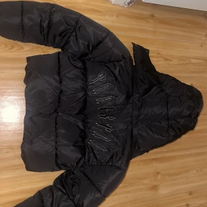 Svart pufferjacka från Trapstar - Svart pufferjacka med avtagbar huva och coolt broderat Trapstar-tryck på ryggen. Jackan har dragkedja framtill, fickor med dragkedja och en rund logga på bröstet. Perfekt för kalla dagar och riktigt street i stilen.