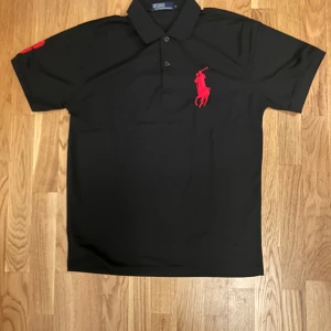 Svart polo t-shirt från Ralph Lauren - Svart polo t-shirt från Ralph Lauren med stor röd broderad logga på bröstet och röda detaljer på ärmen. Klassisk krage med två knappar och korta ärmar. Perfekt för en clean och stilren look.