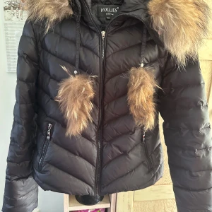 Svart pufferjacka Hollies med pälsdetaljer - Svart pufferjacka från Hollies med snygga pälsdetaljer på huvan och snören. Jackan har dragkedja framtill som saknar hänge, två fickor med dragkedja och quiltad design. Perfekt för kalla dagar när du vill vara både varm och stilren. Äkta päls