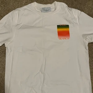 Vit Casablanca t-shirt med färgtryck - Vit t-shirt från Casablanca med färgglatt tryck i grönt, gult och orange på bröstet och ryggen. Stort 'CASA BLANCA'-tryck på ryggen. Klassisk passform och rund hals. Perfekt för dig som gillar stilrena plagg med en twist.